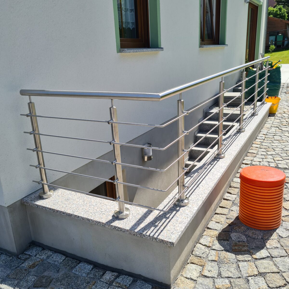 Edelstahl‑Außengeländer mit 5 waagerechten Füllstäben und gebogenem Handlauf an einer Kellertreppe.