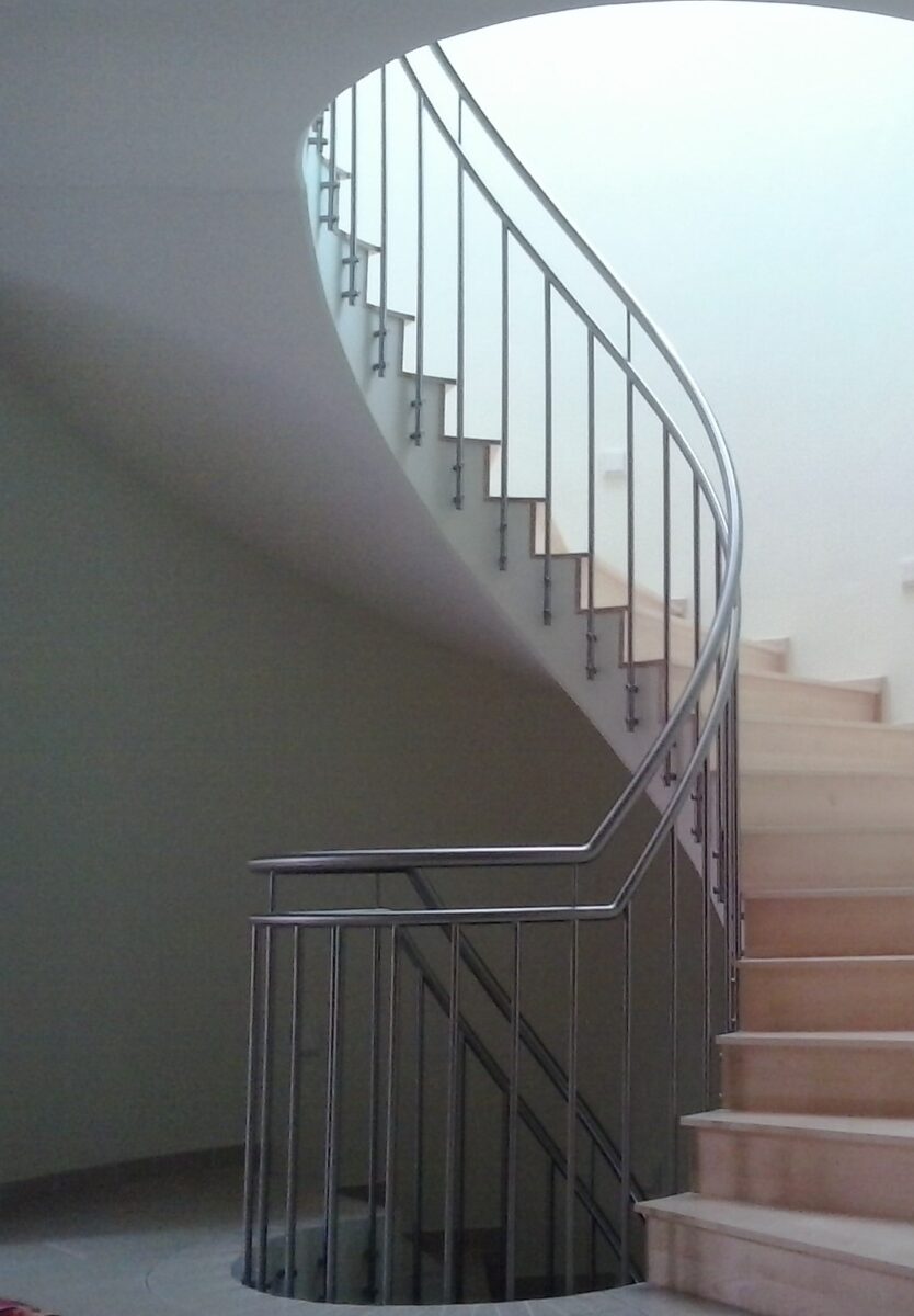 Edelstahl‑Treppengeländer mit senkrechten Füllstäben, an Betonwange verschraubt und dem gewendelten Treppenverlauf folgend gebogen – gefertigt von Edelstahlverarbeitung Schmitt.
